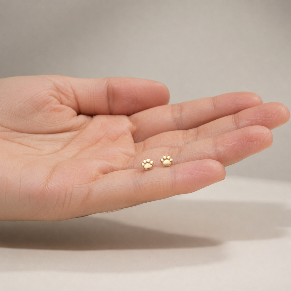 Tiny 14K gold paw print stud earrings displayed in open palm showing scale