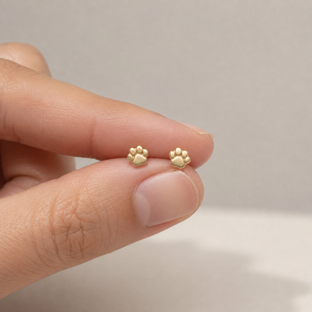 Tiny 14K gold paw print stud earrings held between fingers showing detail and scale